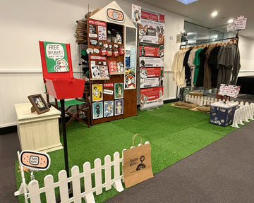 “Home Open” market stall on the Gold Coast—turf and picket fence display with free-stickers mailbox, merch wall, clothing rack, pins and sticker sheets.