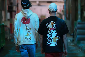 Back view of two individuals wearing 'That's Ruff!' collection apparel—a white hoodie featuring a cat with balloons and a black t-shirt with a bandaged dog graphic. Captured in a moody urban setting, this stylish pairing showcases bold and playful streetwear designs.